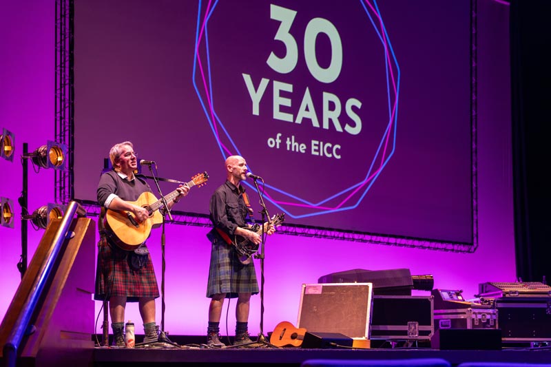 Two musicians with guitars perform on a stage in front of the 30 Years of the EICC logo