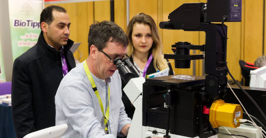 A delegate looks through a microscope as two exhibitors look on